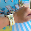 Palm Shore Club silicone Apple Watch band with hand-painted palm trees over soft blue and yellow cabana stripes on an ivory base, shown displayed and worn on Apple Watch against a coastal beach club backdrop.