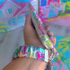 A vibrant La Palma Villa phone scrunchie wrist strap attached to a matching Wavy Luxe phone case with a colorful pink, green, and blue tropical palm tree print.