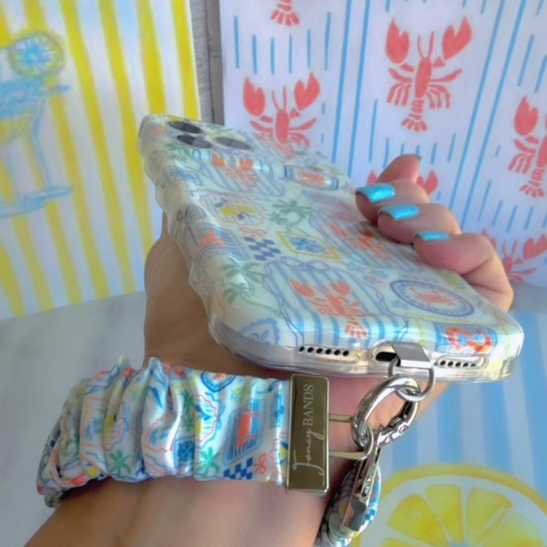A Coastal Catch scrunchie phone wrist strap featuring a blue tile, orange lobster, and yellow lemon pattern attached to a matching Wavy Luxe phone case on a blue and white striped background.