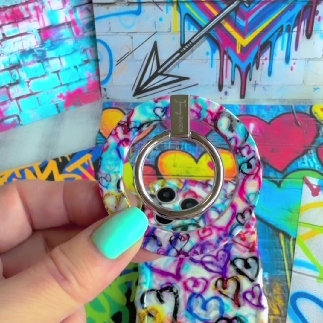 Love Out Loud colorful graffiti heart pattern MagSafe phone grip ring with overlapping neon pink purple and turquoise hearts compatible with iPhone and Samsung