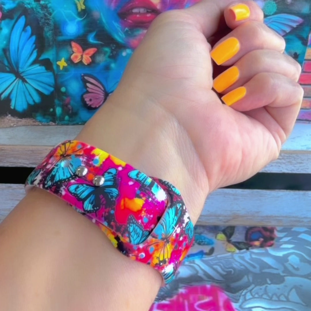Rebel Wings silicone Apple Watch band with graffiti-style butterflies in blue, orange, yellow, and white over a hot pink and magenta paint-splatter background, shown displayed and worn on Apple Watch against a colorful street art butterfly backdrop.